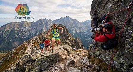 Trail running pe o creastă montană tehnică în România. Un alergător cu bețe (nr. 412) sare peste stânci, surprins de un fotograf agățat pe un perete stâncos. Peisaj spectaculos și logo-ul RunMap.ro în colț.